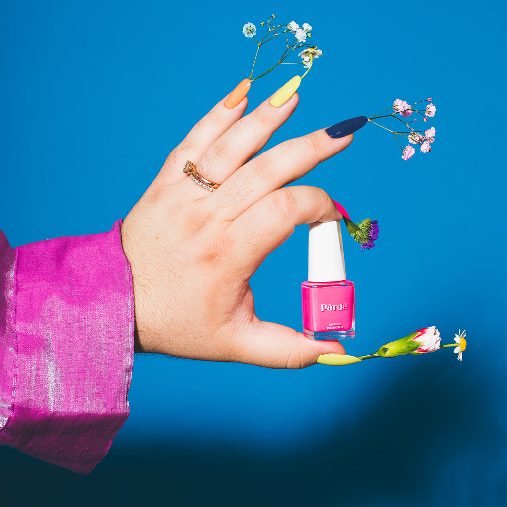 A clear bottle of hot pink nail polish held between two fingers of a hand wearing press-on nails with flowers coming out of them against a blue backdrop.
