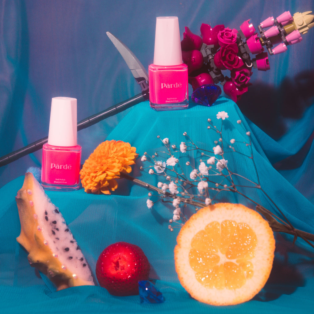 Two clear bottles of hot pink nail polish sitting on a fine blue mesh surrounded by a a slice of dragonfruit and orange, a strawberry, baby's breath and a Lego flower.